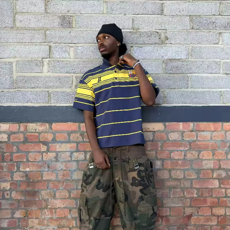 Model wearing the Navy and Yellow striped oversized terrace polo with the FCUL crest, paired with camo cargos and a black beanie.
