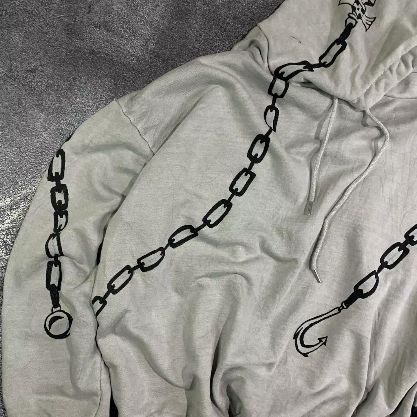 Detailed view of the black chain and hook graphics wrapping around the sleeves and chest of a light grey streetwear hoodie.