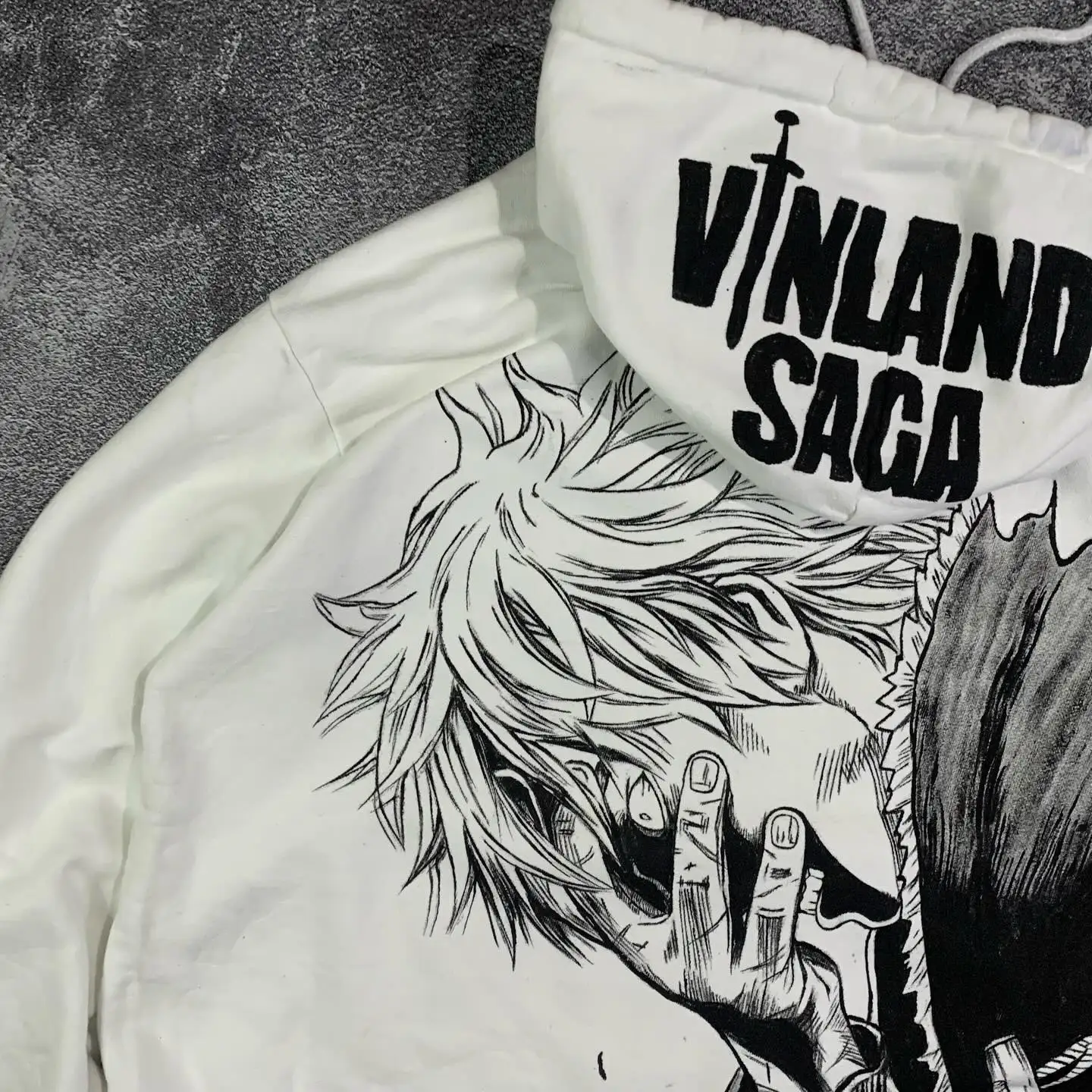 Close-up of the oversized Thorfinn manga graphic and bold Vinland Saga logo printed directly on the hood of a white pullover.